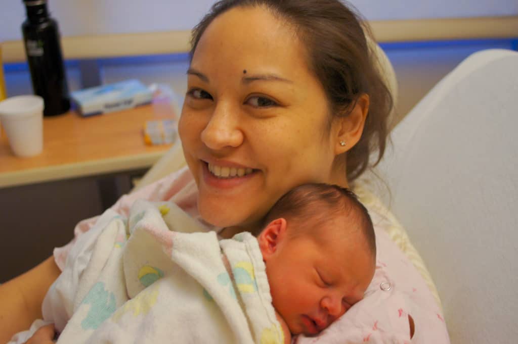 Mother smiling at camera while holding newborn baby in a hospital setting.